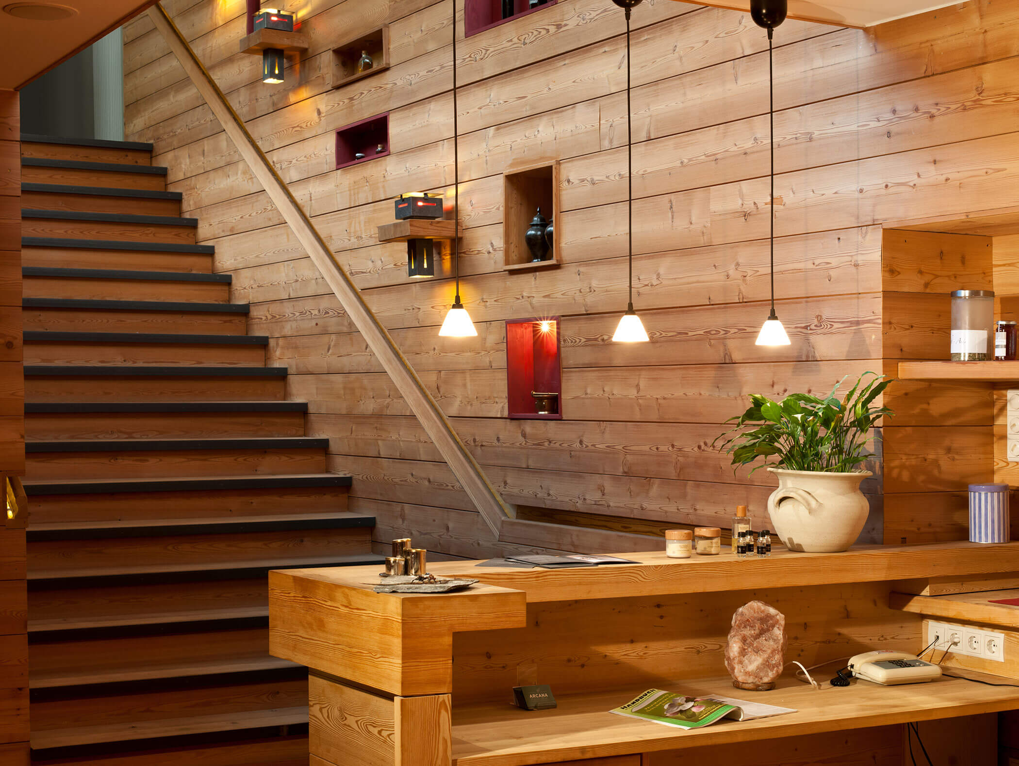 Banco reception e scala in legno, illuminazione calda, pianta in vaso e mensole decorative a parete. - Hotel Moosmair