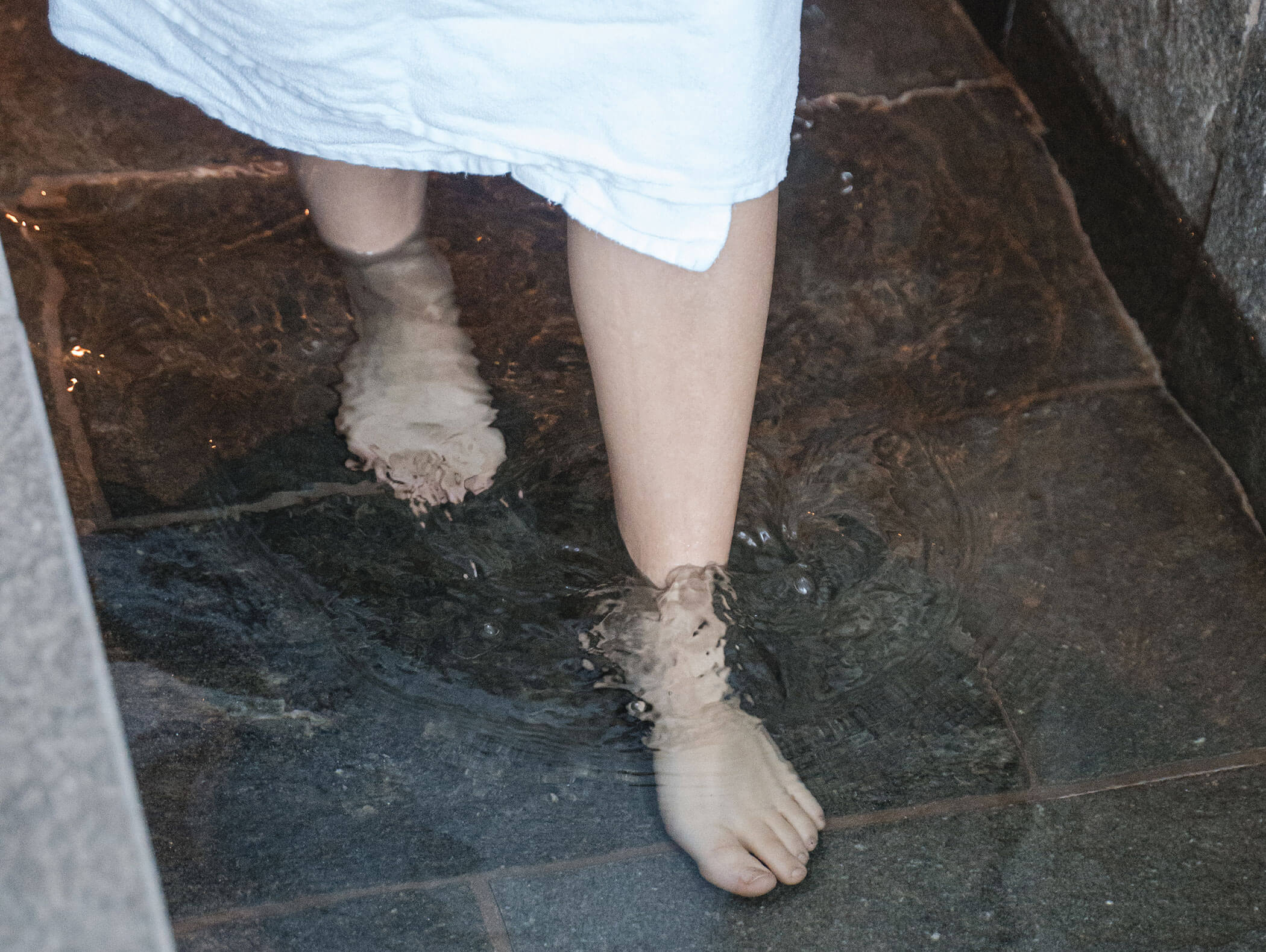 Persona con un asciugamano bianco che sguazza a piedi nudi nell'acqua bassa su piastrelle di pietra. - Hotel Moosmair