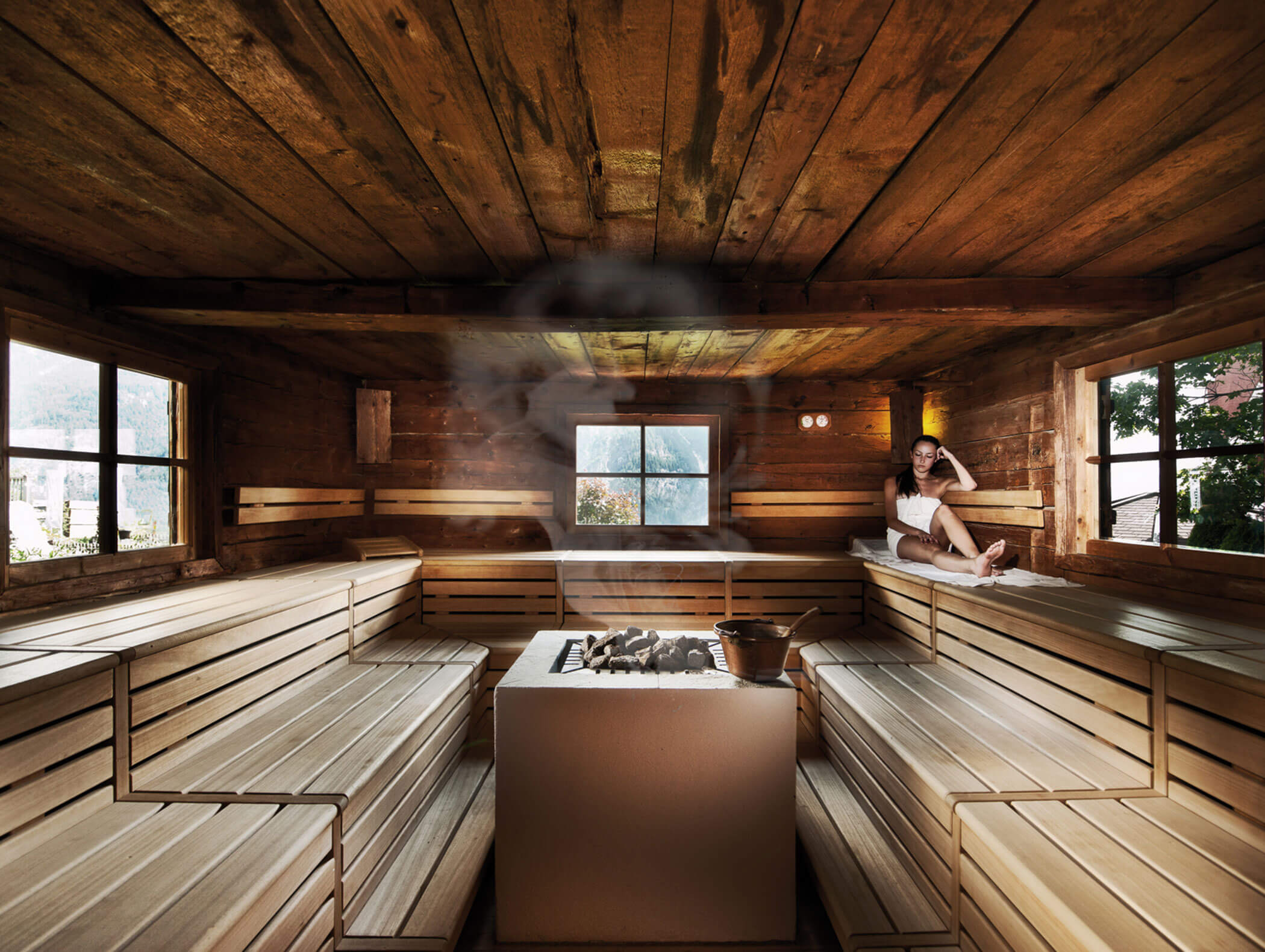 Una donna si rilassa su una panca di legno all'interno di una sauna spaziosa con una stufa centrale in pietra e grandi finestre. - Hotel Moosmair