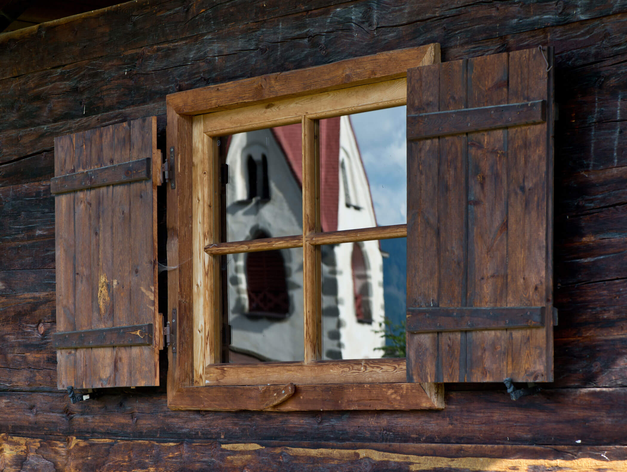 La finestra di una capanna rustica in legno riflette un edificio bianco con tetto rosso e finestre ad arco. - Hotel Moosmair