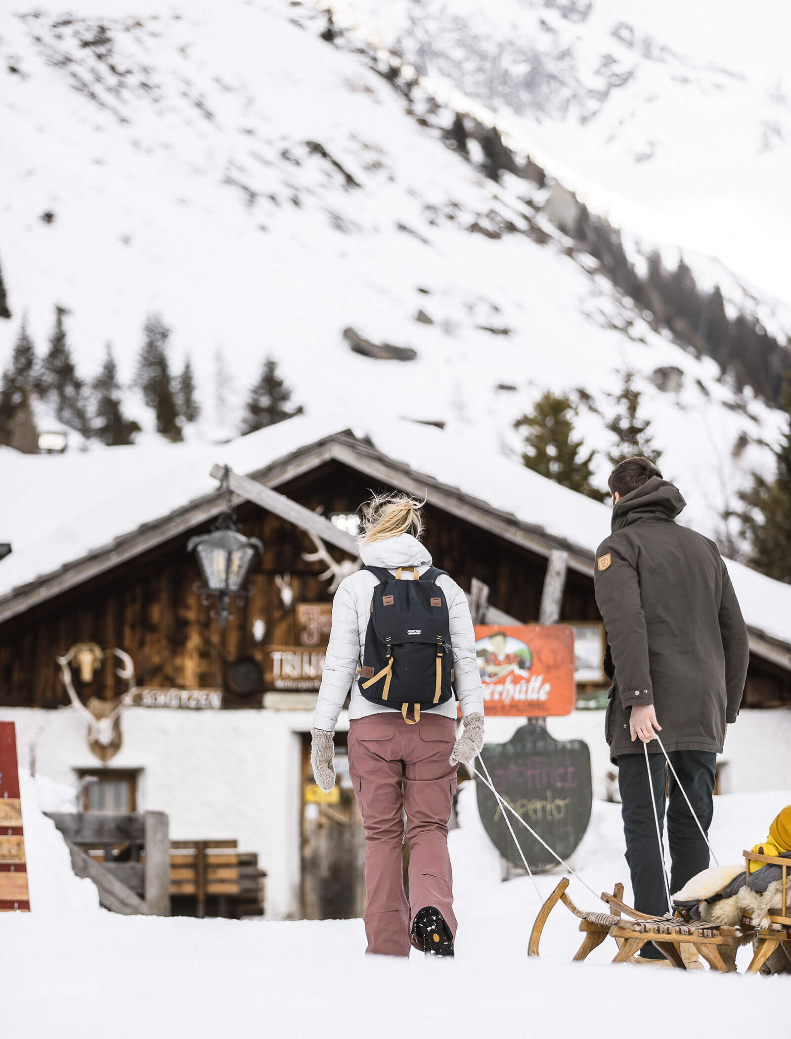 Due persone camminano verso una baita di legno innevata, una delle quali tira una slitta, con montagne e alberi sullo sfondo. - Hotel Moosmair
