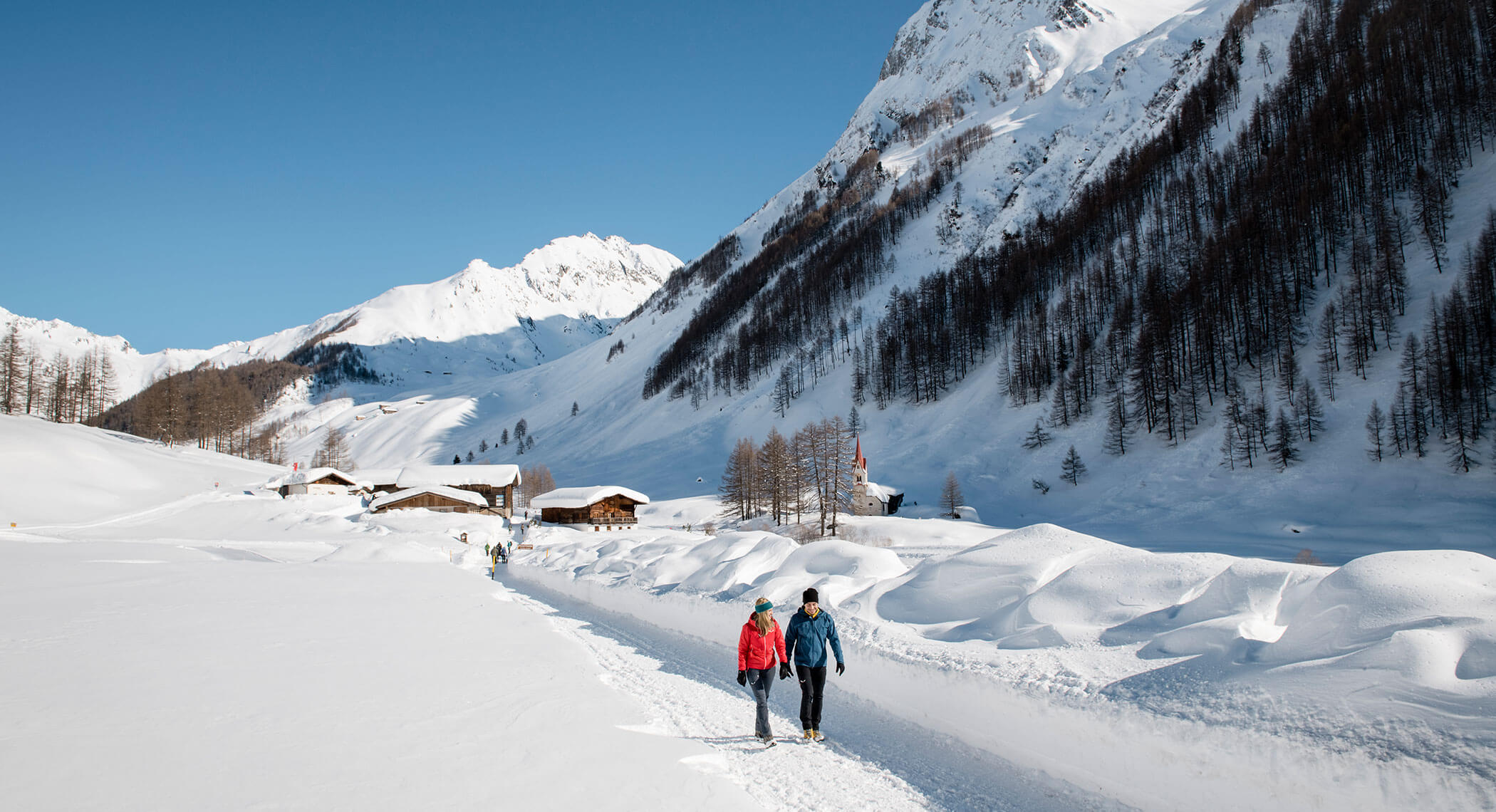 Due persone camminano su un sentiero innevato in un villaggio di montagna sotto un cielo azzurro e limpido. - Hotel Moosmair