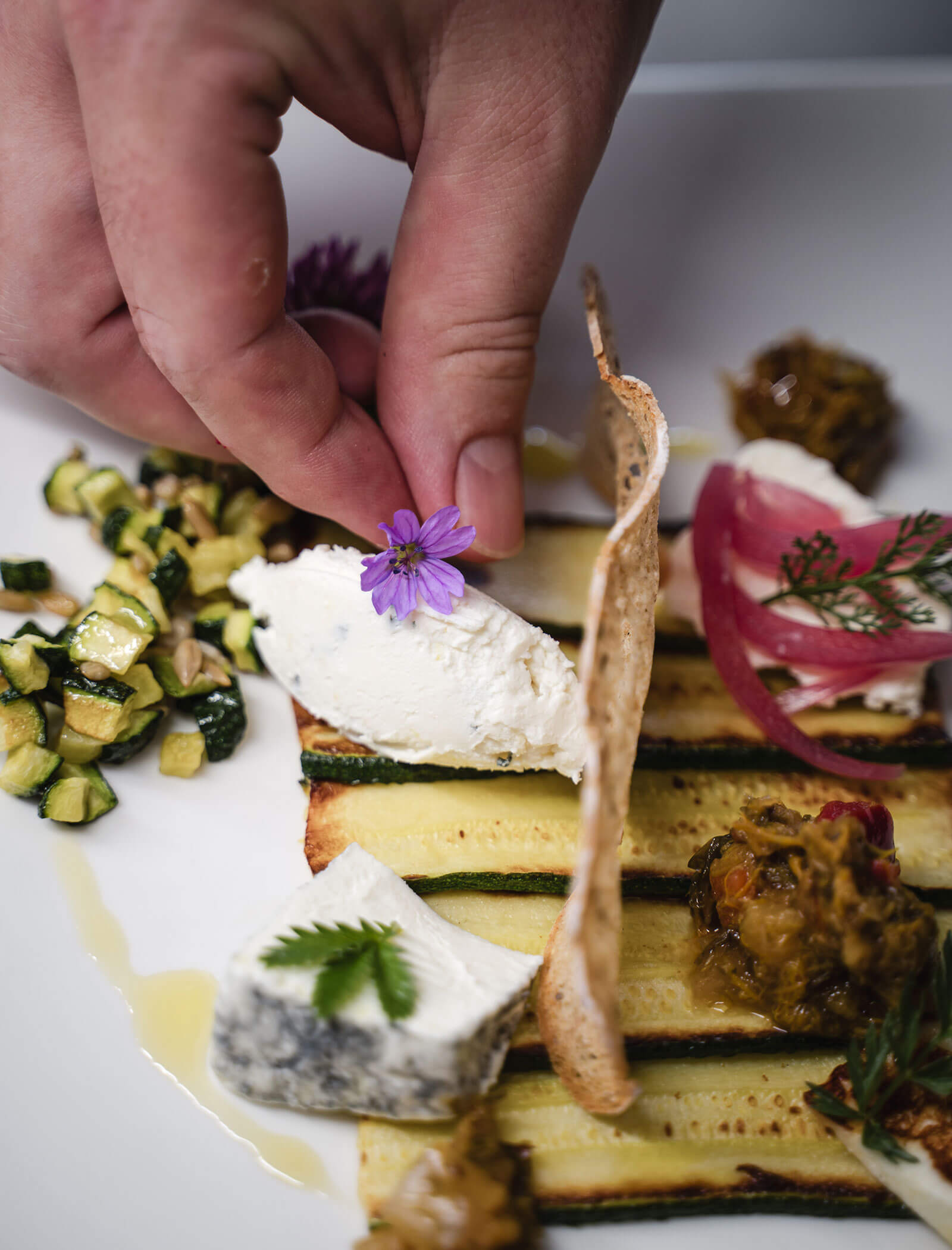 A hand places a purple edible flower on a gourmet plate with cheese, grilled vegetables, and garnish. - Hotel Moosmair