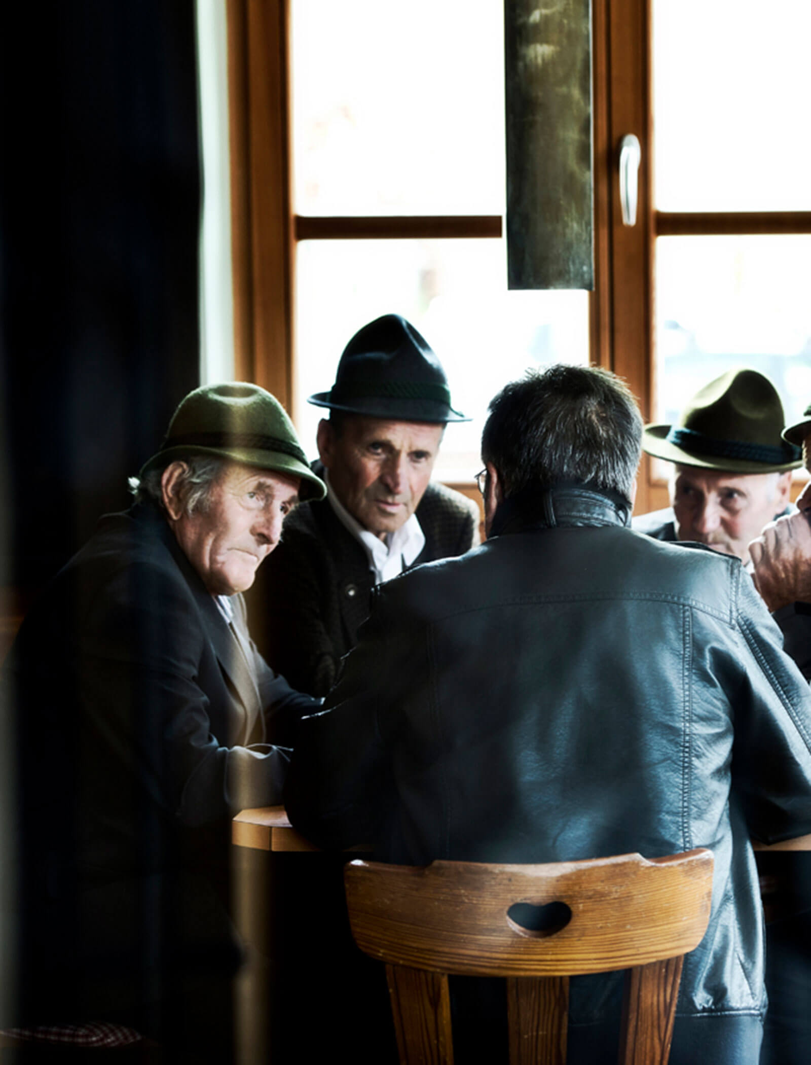 Four older men wearing hats sit around a table, talking, with light coming through a window behind them. - Hotel Moosmair