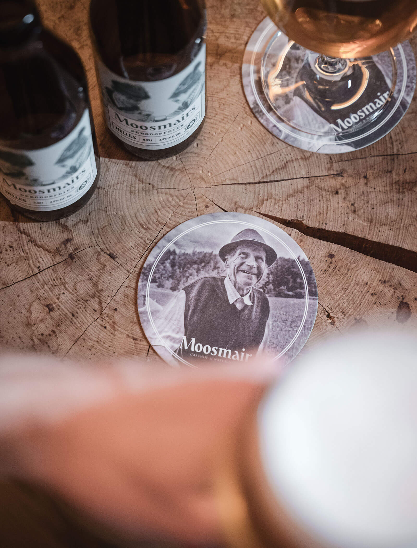 Close-up of beer bottles and a coaster with an older man’s portrait and the text Moosmair on a wooden table. - Hotel Moosmair
