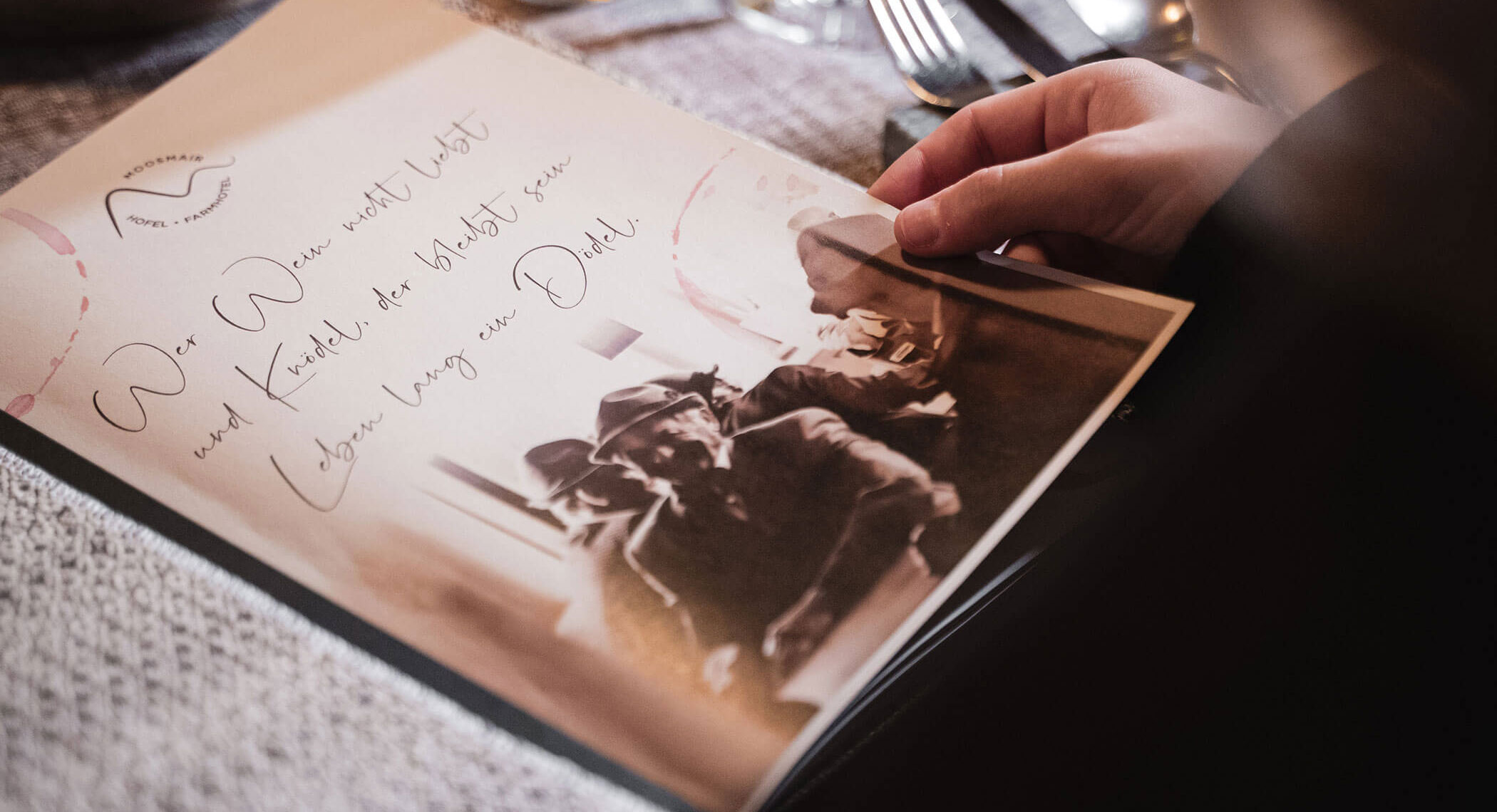 A person holds a menu with German text and a sepia-toned photo of people sitting at a table. - Hotel Moosmair